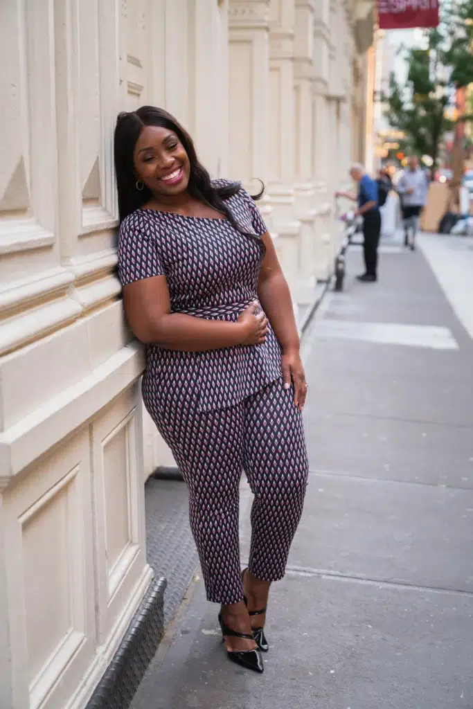Curvy petite woman leaning against a cream ornate building facade on a city sidewalk, wearing a matching navy, pink, and white diamond geometric print co-ord set with a bardot-style short-sleeve top and cropped ankle pants, styled with black patent pointed-toe heeled pumps with an ankle strap, simple silver hoop earrings, and a bold pink lip.