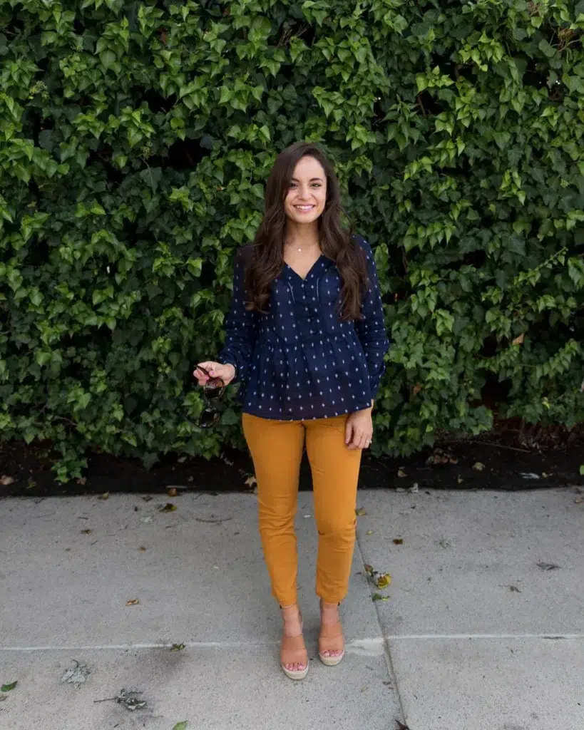 Petite woman in a navy blue small-dot printed peasant blouse with a V-neckline and long sleeves, paired with slim-fit mustard yellow ankle pants and nude espadrille wedge sandals, standing on a sidewalk in front of a lush green ivy-covered wall, holding a pair of sunglasses.