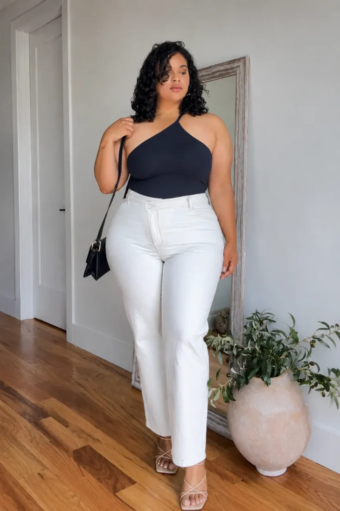 Petite plus-size woman with natural curly dark hair wearing a navy angular halter neck bodysuit tucked into high-rise crisp white straight-leg jeans with a subtle flare at the hem, carrying a small structured black leather crossbody bag with a top handle and gold clasp hardware, wearing nude strappy lace-up flat sandals, standing in front of a large floor-length mirror with a distressed gray wood frame, beside a large round beige ceramic vase holding olive branches, on warm hardwood floors against a light gray wall.