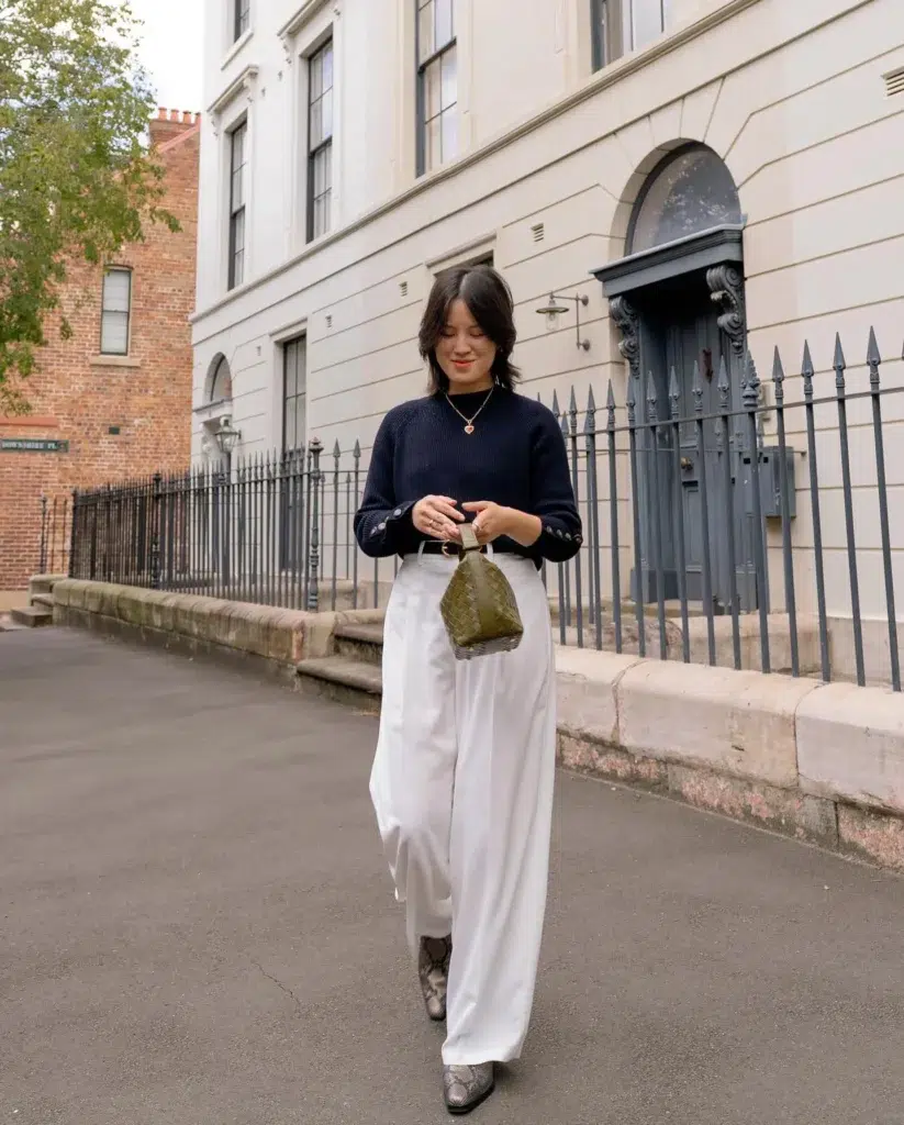 Petite woman walking down a city street, wearing a navy ribbed mock-neck knit with button cuffs tucked into tailored white wide-leg trousers, snake-print pointed boots, and holding a small olive woven bag, with a gold heart pendant, cream building and iron fence behind her.