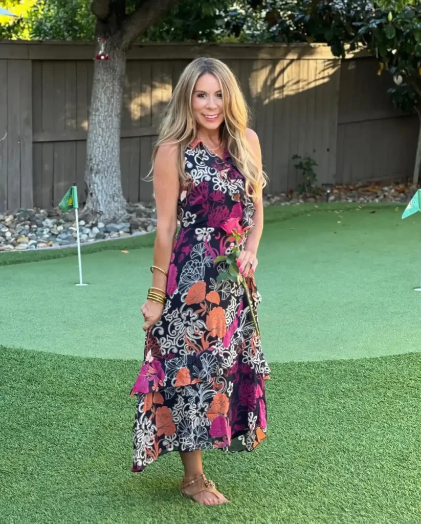 Woman in a dark navy sleeveless midi dress with large bold fuchsia, orange, and white botanical scroll florals, a fitted bodice, and tiered ruffle hem, wearing flat tan strappy sandals with logo buckle hardware and stacked gold bangle bracelets, holding a pink rose, standing on a backyard putting green with a large tree and wooden fence in the background.