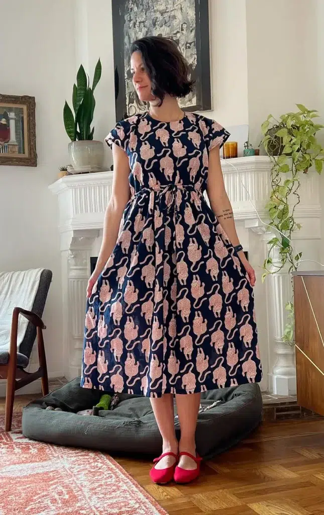 Petite woman in a dark navy and pink novelty print midi dress with cap sleeves, a drawstring-cinched waist, and a full gathered skirt, wearing red Mary Jane flats, standing in a bright living room with mid-century modern furniture and indoor plants.