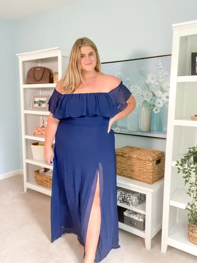 Petite plus-size woman with long straight blonde hair wearing a floor-length navy blue off-shoulder chiffon maxi dress with a layered ruffle flounce at the neckline and a front thigh-high slit, standing barefoot indoors with one hand on her hip in a confident pose, in front of a white bookshelf styled with wicker baskets and a brown leather handbag, with a large floral canvas painting and a second white shelving unit visible in the background against a soft blue-gray wall.