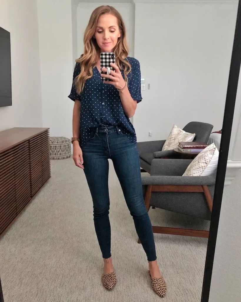 Petite woman in a navy blue polka dot short-sleeve button-front blouse with a front tuck, paired with dark indigo high-rise skinny jeans with a raw frayed ankle hem and tan pointed-toe cheetah print mule flats, taking a mirror selfie in a bright modern home living room.