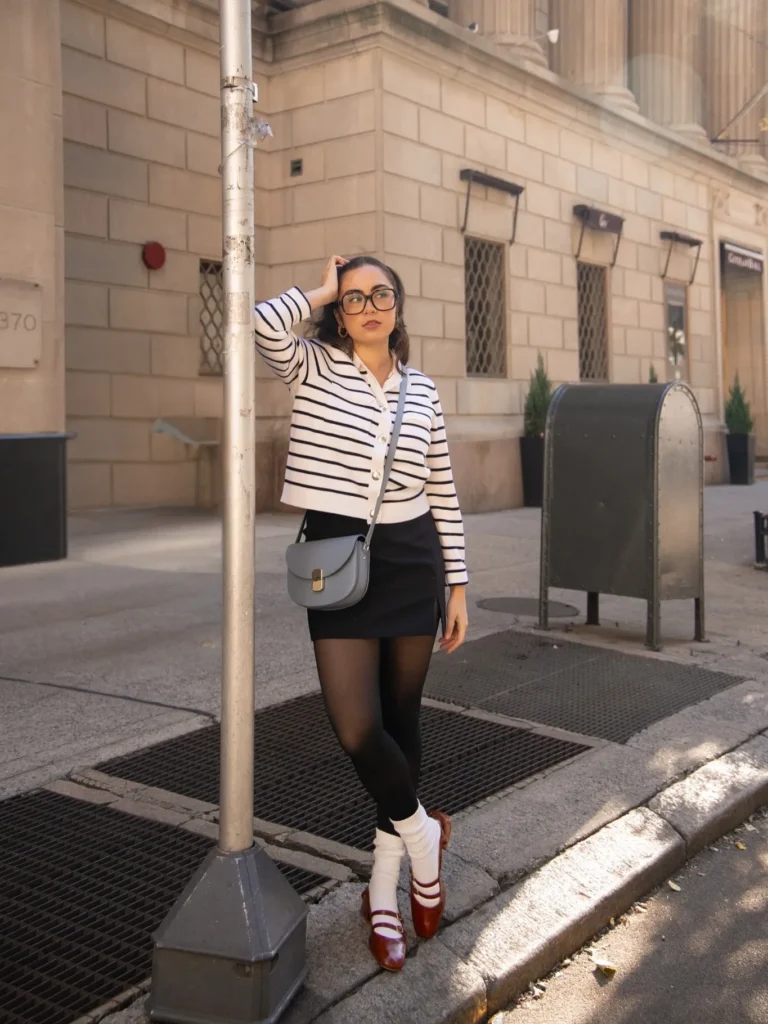 Petite woman in a cropped white and navy striped buttoned cardigan layered over a white collared shirt, paired with a black mini skirt, black sheer tights, and white slouchy crew socks worn over the tights, wearing dark burgundy patent leather double-strap Mary Jane flats, carrying a dusty blue-gray structured saddle crossbody bag with a gold clasp, standing on a city sidewalk beside a lamp post with a stone building in the background.