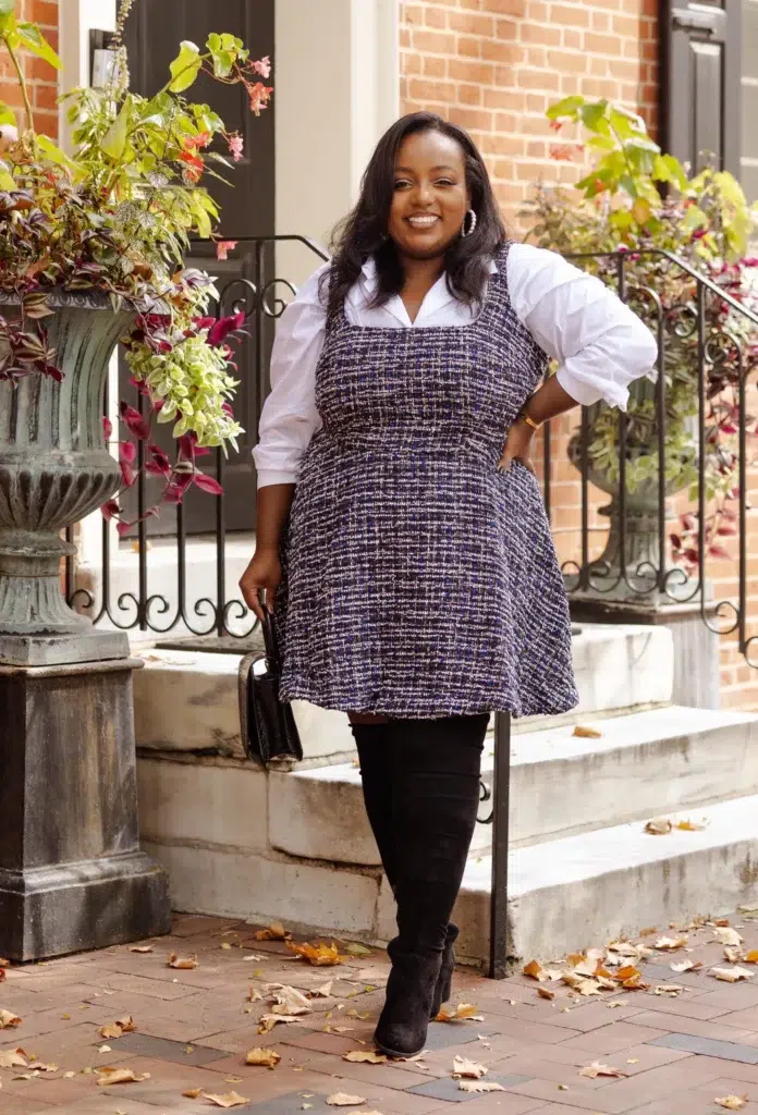 Petite plus-size woman with long dark wavy hair wearing a navy and lavender multicolor boucle tweed A-line pinafore dress layered over a crisp white poplin button-front shirt with voluminous puffed balloon sleeves, paired with black skinny pants and black suede over-the-knee heeled boots, holding a small black structured clutch, wearing large white statement hoop earrings and a gold chain bracelet, standing confidently on a brick sidewalk in front of a brownstone with ornate iron railings and large stone planters filled with colorful autumn foliage.