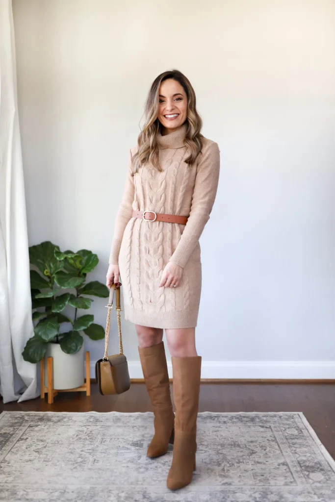 Petite brunette woman in an oatmeal cable knit turtleneck mini sweater dress cinched at the natural waist with a tan leather belt with a gold buckle, paired with tall tan suede knee-high boots and a structured two-tone olive and tan handbag with a chain strap, standing on a vintage rug against a clean white wall with a large fiddle leaf fig plant in the background.