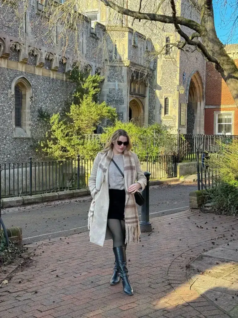 Petite woman with loose blonde wavy hair walking on a brick pathway in front of a large Gothic stone church or cathedral with iron railings and green hedges, wearing a long open oatmeal cream wool-blend coat with a very large oversized plaid blanket scarf in cream and camel brown check with fringe ends draped over both shoulders and the coat, a light grey soft knit visible at the chest, a structured black mini skirt, dark charcoal semi-opaque tights, classic tall black leather equestrian knee-high boots with a modest heel, dark cat-eye sunglasses, and a small black crossbody saddle bag at her hip.