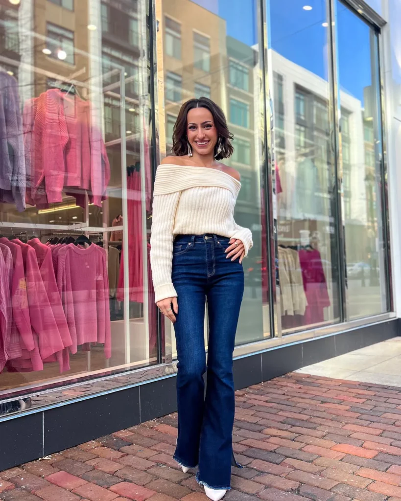 Petite woman in dark indigo high-rise flare jeans with a raw frayed hem, wearing a chunky cream ribbed off-the-shoulder sweater with a wide fold-over neckline draped across both shoulders, styled with white pointed-toe heels and large statement drop earrings on a brick city sidewalk.
