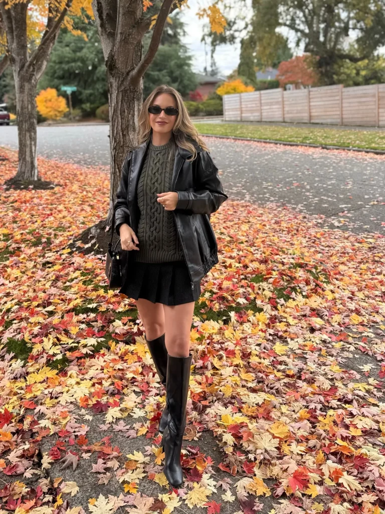 Petite woman in an oversized black leather zip-front moto jacket worn fully open over a deep olive army-green chunky cable knit crewneck sweater, paired with a black pleated mini skirt and tall black flat-heeled knee-high leather boots, carrying a small dark structured bag at her side, wearing small black rectangular sunglasses, with long blonde wavy hair worn down, standing on a suburban street beside a bare-trunked tree on an overcast fall day with a vibrant full carpet of red, orange, and gold fallen maple leaves covering the ground and surrounding trees showing peak autumn color.