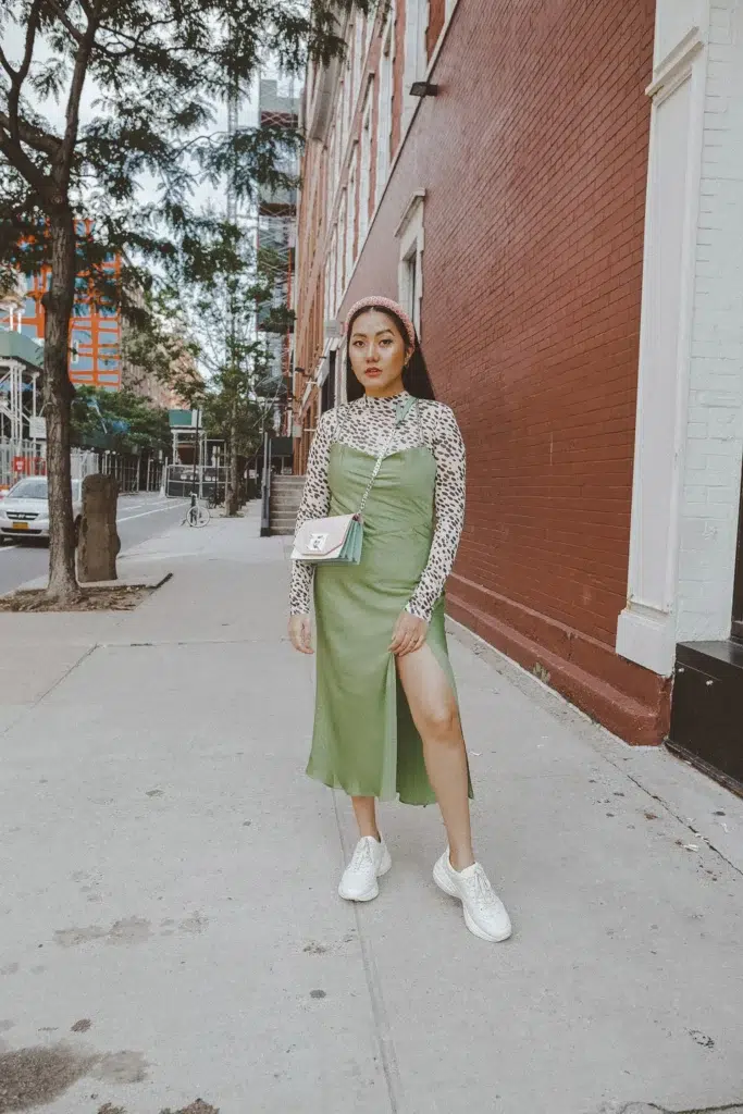 Petite East Asian woman with long straight dark hair and a dusty pink textured padded headband standing on a city sidewalk in front of a red brick building, wearing an olive sage green satin midi slip dress with a side slit layered over a long-sleeve muted neutral leopard animal print top, paired with white chunky lace-up sneakers and a small blush pink structured crossbody bag on a chain strap, with small gold earrings.