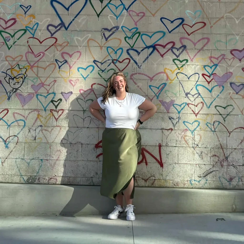 Curvy petite woman standing against a concrete wall covered in colorful spray-painted heart outlines in rainbow colors, wearing a fitted white crew-neck t-shirt tucked into an olive green satin wrap midi skirt with an asymmetric tulip hem, paired with white Nike court sneakers, layered gold and silver necklaces, and a black smartwatch, with shoulder-length blonde hair worn loose.