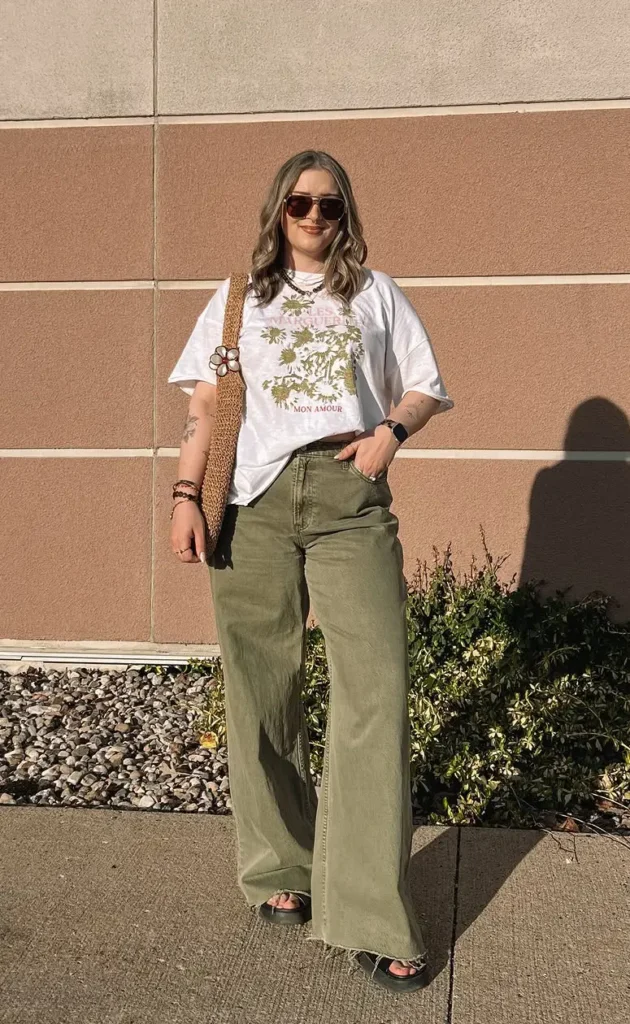 Petite woman in faded olive wide leg jeans, an oversized white daisy graphic tee with a front tuck, black chunky slide sandals, and a woven crossbody bag, photographed outdoors