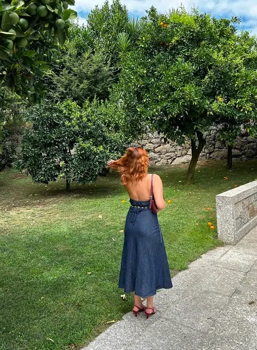 Woman photographed from behind wearing a dark navy denim backless midi dress with thin spaghetti straps, a tied or belted waist detail, and a soft flared A-line skirt, styled with a small wine-burgundy crossbody bag worn across the back and deep red pointed-toe kitten heel mules, standing on a stone garden path surrounded by lush green citrus trees with oranges visible in the branches and a stone wall backdrop.