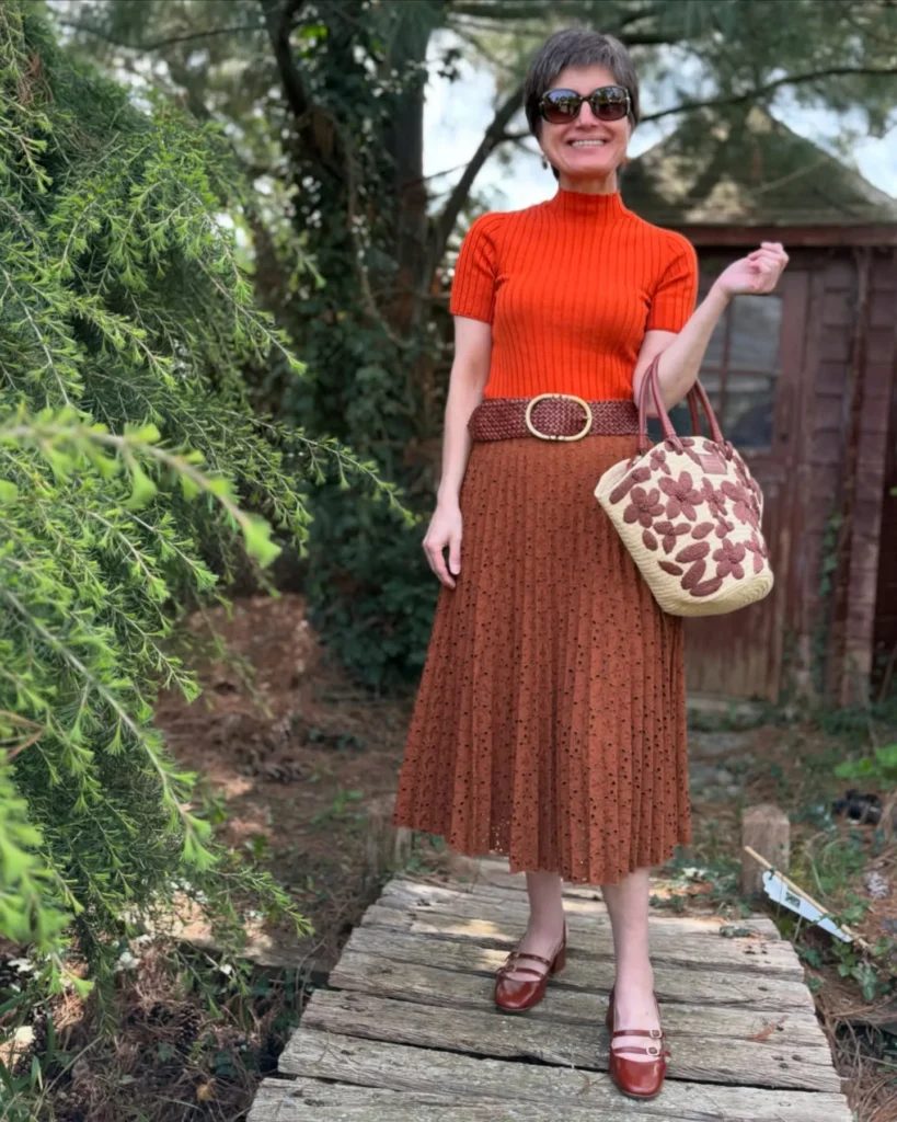 Petite woman with short hair standing on a wooden garden walkway, wearing a bright orange ribbed mock-neck short-sleeve knit tucked into a rust eyelet midi skirt with a woven belt, brown Mary Jane flats, and carrying a straw tote with floral detail, greenery behind her.
