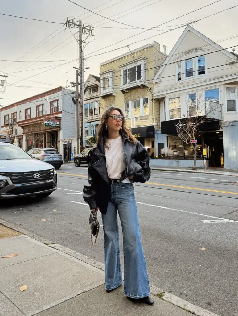 Petite woman in very light wash super wide-bell flare jeans wearing an oversized open black leather jacket over a tucked white basic tee, styled with a black decorative-buckle belt, black pointed-toe flat boots, clear-frame glasses, and a small black quilted chain bag held in hand on a city street.