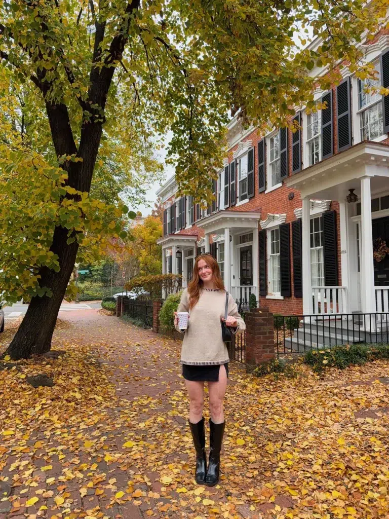 Petite woman with long red hair wearing an oversized oatmeal knit sweater with dark contrast trim at the neckline and hem, layered over a black mini skirt barely visible at the hem, paired with tall structured black knee-high boots with a block heel and square toe, carrying a black shoulder bag, holding a holiday-printed paper coffee cup, standing on a leaf-covered brick sidewalk in front of classic red brick colonial townhouses with black shutters and white trim on an overcast autumn day.