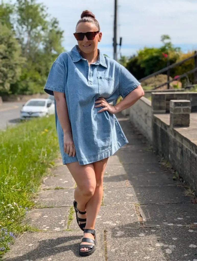 Woman in a light blue wash oversized short-sleeve polo-collar denim mini dress with a partial button placket, a small chest patch pocket, and wide boxy sleeves, styled with tortoiseshell rectangular cat-eye sunglasses, hair up in a high bun, and black chunky triple-strap platform sandals, standing on a sunny suburban pavement with green trees and a low stone wall in the background.