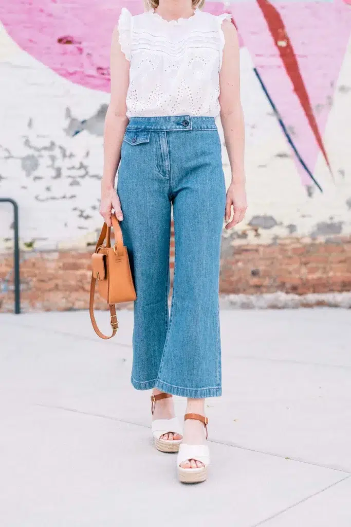 Petite woman wearing high-rise wide-leg cropped jeans with a white eyelet blouse, wedge sandals, and tan handbag.