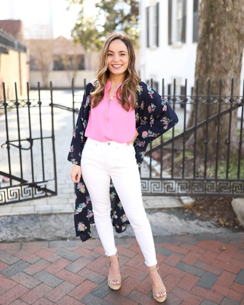Petite woman standing on a brick sidewalk by an iron fence, wearing a hot-pink henley blouse tucked into high-waisted cropped white skinny jeans, an open navy floral duster kimono, and nude espadrille heels, with a gold pendant necklace.