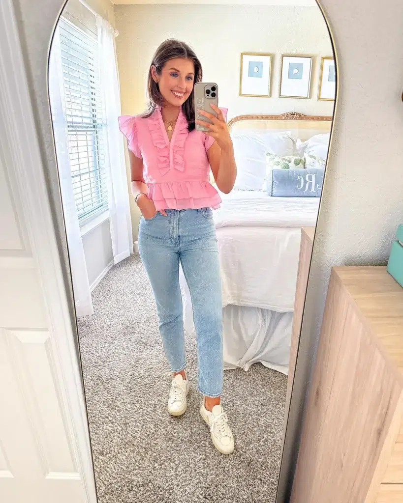 Petite woman taking a mirror selfie in a bedroom, wearing a bubblegum-pink ruffle peplum top with flutter sleeves, high-rise light-wash cropped straight jeans, and white sneakers, with a gold pendant necklace, standing in front of a bed and wood dresser.