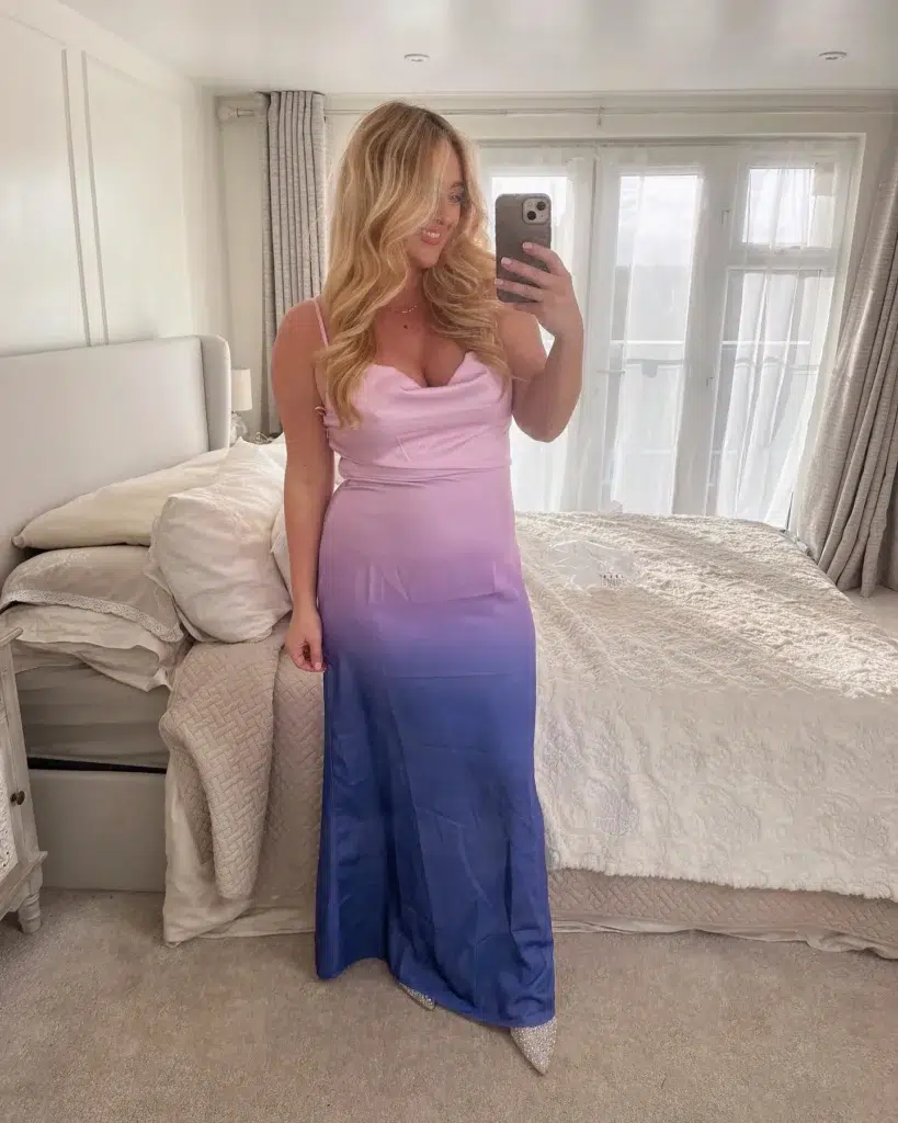 Curvy petite woman taking a bedroom mirror selfie in a floor-length ombre satin maxi dress transitioning from soft blush pink at the cowl neckline through lavender to deep cobalt blue at the hem, styled with crystal-embellished pointed-toe heels, a delicate gold necklace, and voluminous blonde wavy hair, standing in a grey and white bedroom.