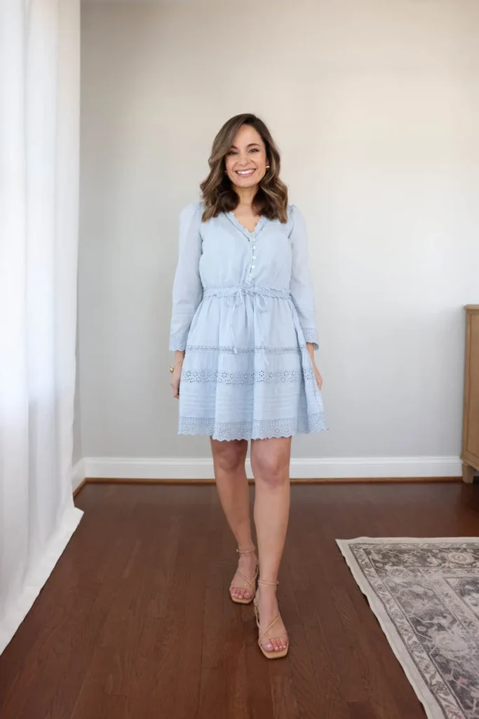 Petite woman standing in a bright room, wearing a powder-blue eyelet-trimmed mini dress with long sleeves, a button front, and a drawstring waist, paired with skin-tone strappy heeled sandals, with a patterned rug and wood dresser behind her.