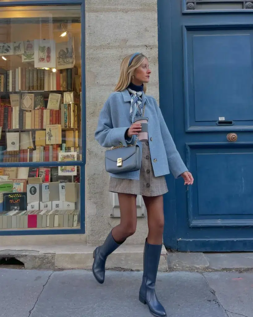 Petite woman with straight blonde hair and a blue fabric headband standing on a Parisian pavement beside a deep blue painted bookshop door and a warm-lit antiquarian bookshop window, wearing a powder blue oversized wool jacket with gold button details over a navy ribbed turtleneck, a blue and grey printed silk scarf tied loosely at the collar, a grey and taupe plaid button-front mini skirt, bare legs, slate blue tall flat knee-high leather equestrian-style boots, gold clip earrings, and carrying a structured blue crossbody bag with a gold chain strap and gold clasp, holding a coffee cup.