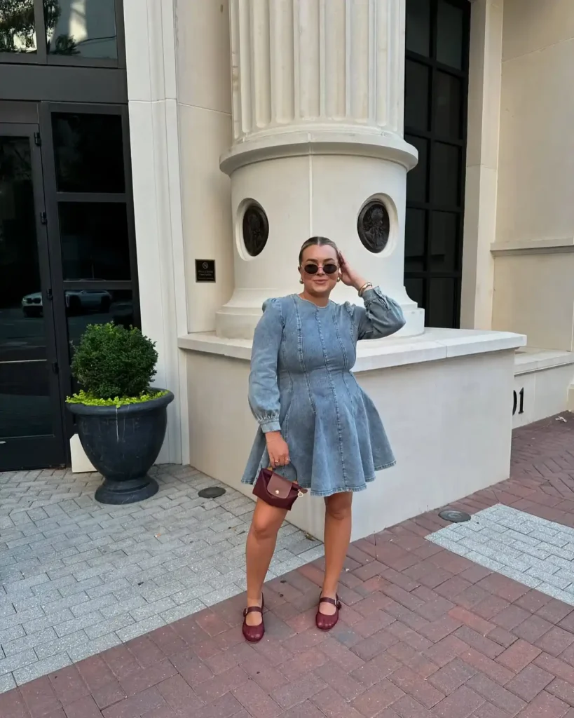 Woman in a light blue wash long-sleeve fit-and-flare denim mini dress with bishop puff sleeves and gathered cuffs, a round neckline, fitted vertical-seamed bodice, and a short flared skater skirt, styled with deep burgundy leather Mary Jane flats with a single buckle strap, a small structured burgundy micro-bag with a gold clasp, small oval tinted sunglasses, gold hoop earrings, and a gold bracelet, with hair slicked back, standing on a brick-paved outdoor area beside a white classical stone column building with a black door and dark urn topiary planter.