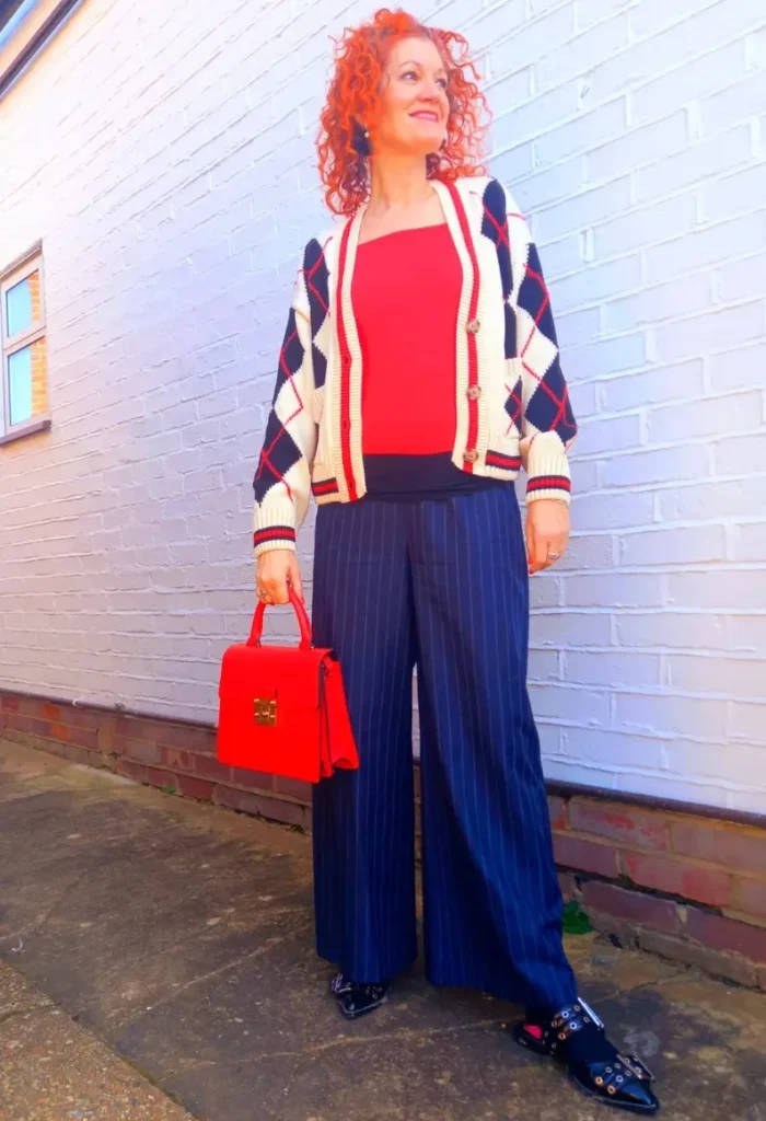 Woman with voluminous red curly hair standing outdoors against a white brick wall, wearing a cream, navy and red argyle cropped cardigan open over a fitted red top, navy pinstripe wide-leg palazzo trousers, and black pointed-toe buckle-detail flat shoes, holding a structured red top-handle bag with gold hardware.