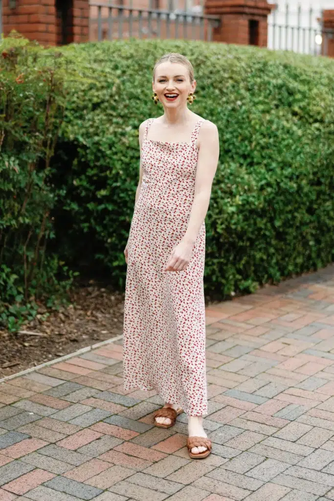 Tall slender woman with blonde hair pulled back sleekly, wearing a white midi slip dress with a small scattered red floral ditsy print and wide square straps, paired with tan woven flat slide sandals, large gold cluster statement earrings, and a bold red lip, standing and laughing on a herringbone brick pathway in front of a trimmed green hedge with red brick buildings behind her.