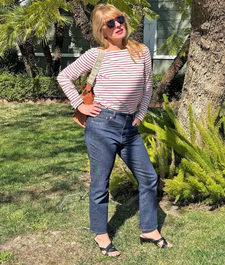 Petite woman standing on a lawn in front of trees wearing high-waisted dark indigo straight leg cropped jeans, a long-sleeve white and red horizontal stripe Breton boat-neck top worn untucked, black strappy low-heeled mule sandals, and a tan camel leather crossbody bag with a woven logo-print canvas shoulder strap, with tortoiseshell sunglasses and a red lip.
