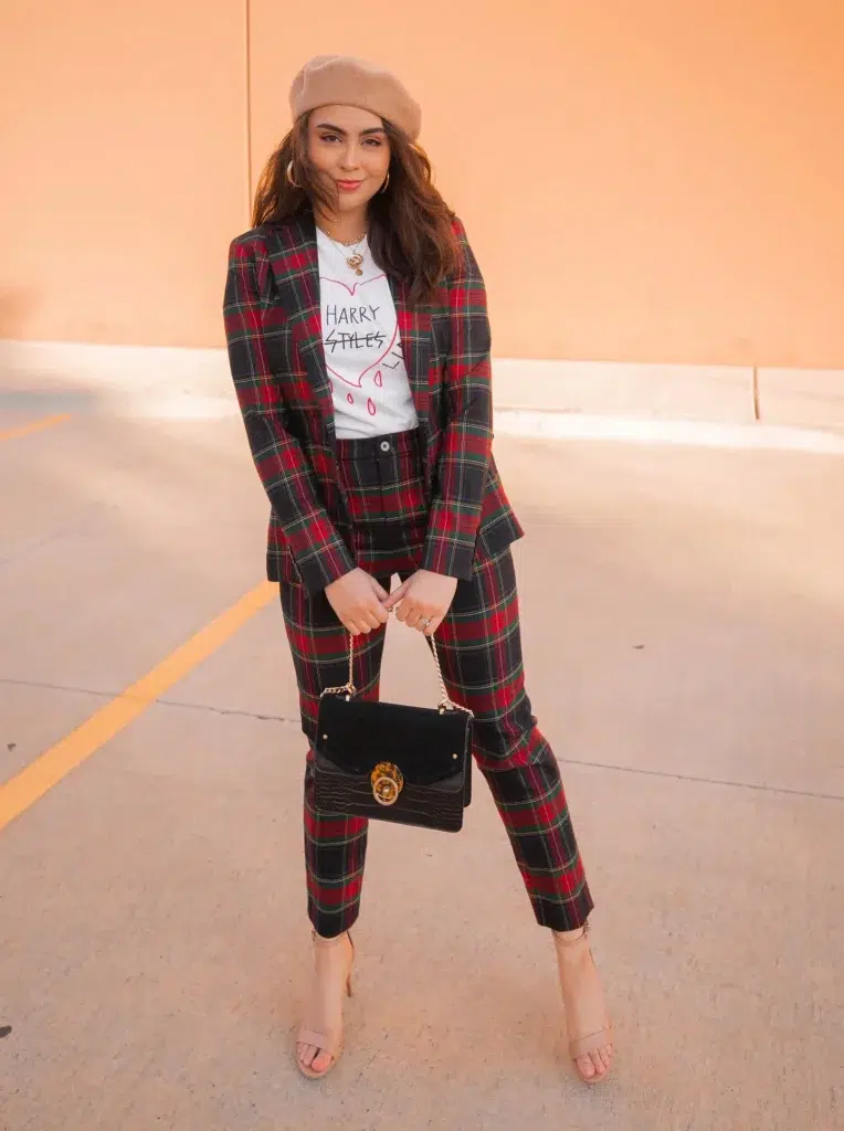 Petite woman in a matching dark tartan plaid suit set with a single-button blazer and cropped slim-fit trousers in a navy, red, and green plaid pattern, worn open over a white graphic print t-shirt, paired with nude strappy heeled sandals and a camel wool beret, carrying a black structured chain-strap bag with a tortoiseshell clasp detail, wearing layered gold necklaces and gold hoop earrings, standing against a warm peach-toned exterior wall.