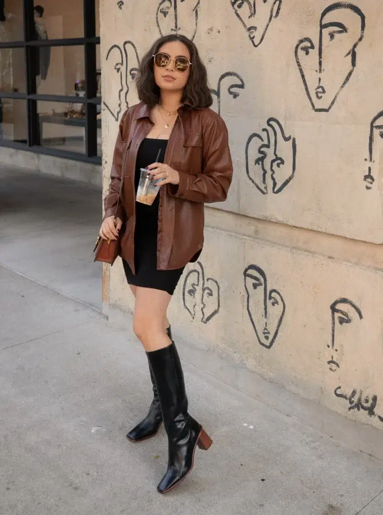 Petite woman in an oversized rich brown leather button-front shacket worn fully open over a fitted black ribbed bodycon mini dress, paired with tall black knee-high boots with a stacked block wooden heel and square toe, carrying a small structured brown boxy bag held at the side, wearing round gold-frame sunglasses with amber lenses and layered delicate gold necklaces, holding an iced coffee, standing on a city sidewalk beside a beige wall covered in abstract black line-drawn face illustrations.