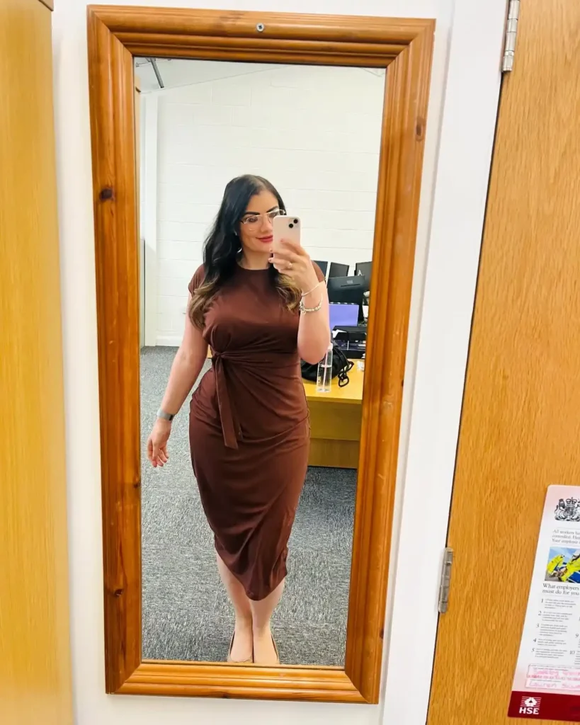 Petite woman with long dark wavy hair and glasses in a rich chocolate brown short-sleeve round-neck fitted midi dress with a side fabric knot detail at the natural waist, paired with nude flat shoes, taking a mirror selfie in an office setting.