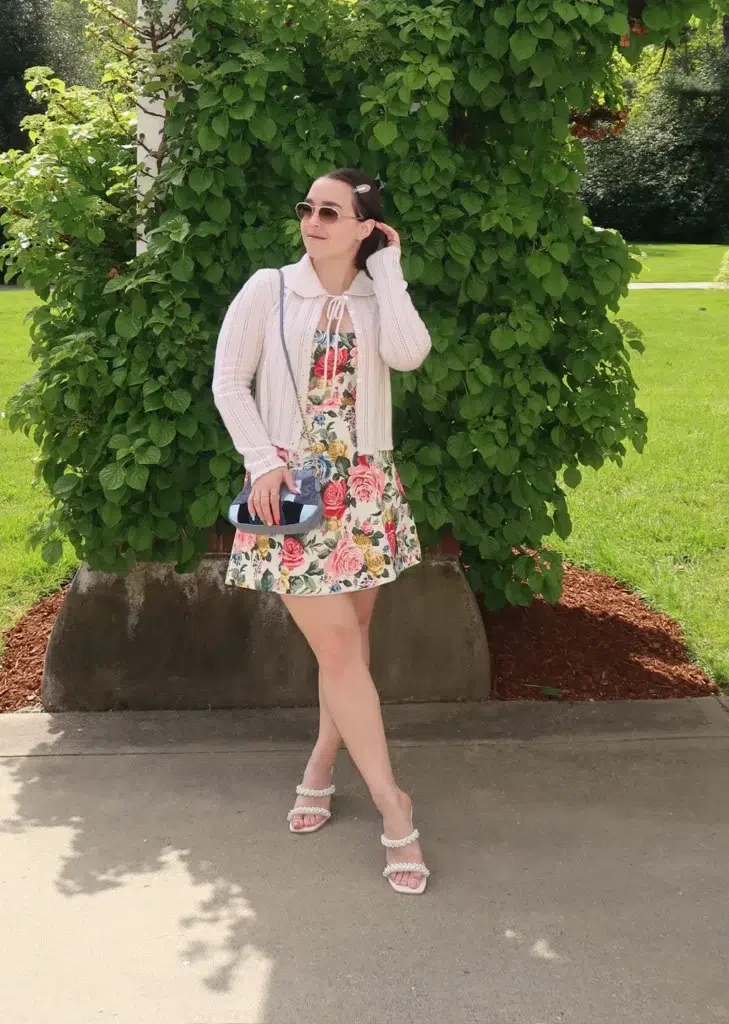 Petite woman standing on a garden path under a white pergola, wearing a pastel rose-print mini dress layered under a cropped white pointelle cardigan with an oversized collar, pearl-strap heeled sandals, sunglasses, and a hair clip, holding a small bag, with green vines behind her.