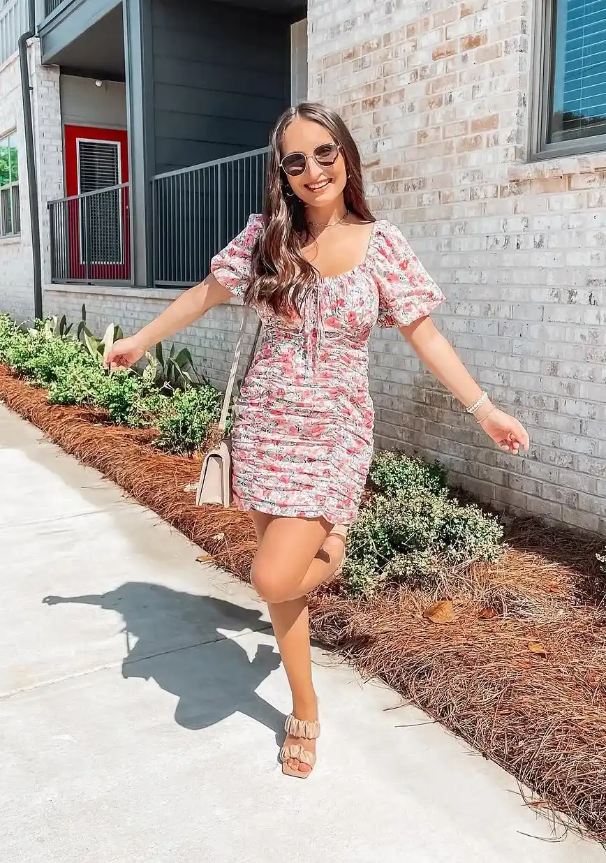 Petite woman in a blush pink and white floral ruched mini dress with short puff sleeves and a drawstring tie neckline, wearing nude ruched flat sandals and carrying a small structured blush shoulder bag, with round sunglasses and layered delicate necklaces, walking on a sunny sidewalk outside a brick apartment building.
