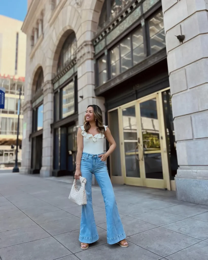 Petite woman in very light wash super wide-bell flare jeans with a double-stitched visible hem, wearing a fitted cream ruffled tank top with a sweetheart neckline and ruffle shoulder straps, paired with champagne heeled sandals and a small white quilted structured mini bag on a city sidewalk.