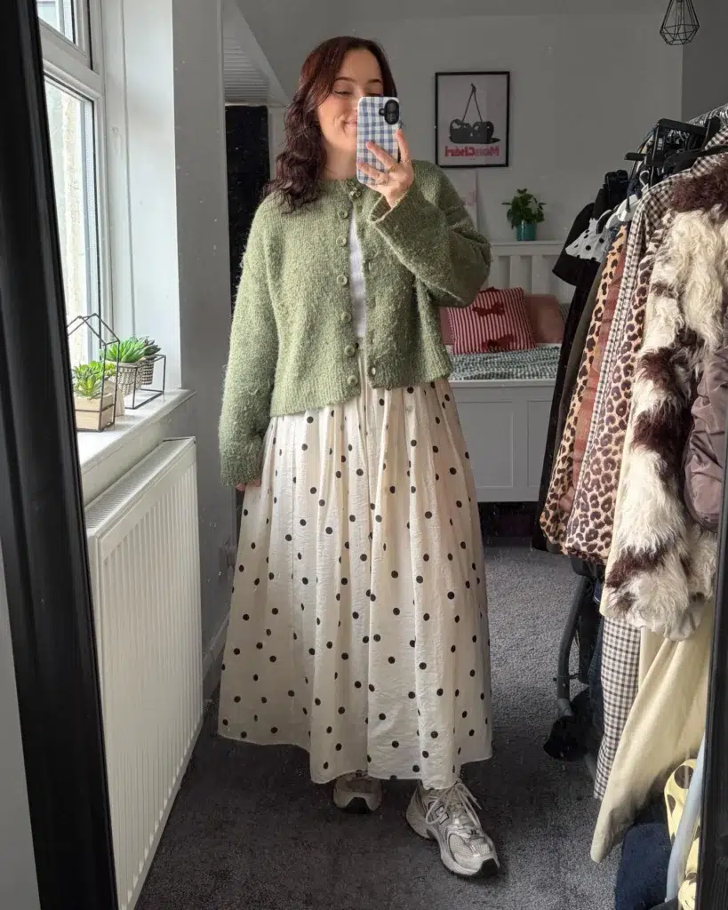 Petite Woman with long dark reddish-brown wavy hair taking a mirror selfie in a bright bedroom, wearing a sage green fluffy bouclé cropped button-front cardigan with round buttons over a white tee, a cream gathered maxi skirt with large dark polka dots in a crinkled fabric, and chunky silver retro sneakers just visible below the full skirt hem, with a clothing rack with mixed prints and a window with small succulents in the background.