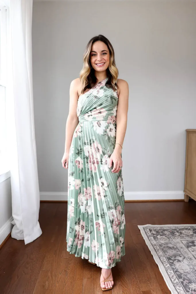 Woman wearing a sage green and dusty pink floral pleated maxi dress with a one-shoulder ruched bodice, paired with tan strappy heeled sandals.