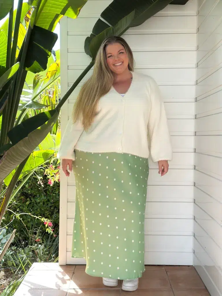 Petite plus-size woman with long straight blonde hair wearing a cream fuzzy cropped V-neck button-front cardigan over a sage green polka dot flowy maxi skirt with white dots, layered over a white camisole, with white chunky platform sneakers, standing in front of a white shiplap wall with large tropical banana leaves to one side.