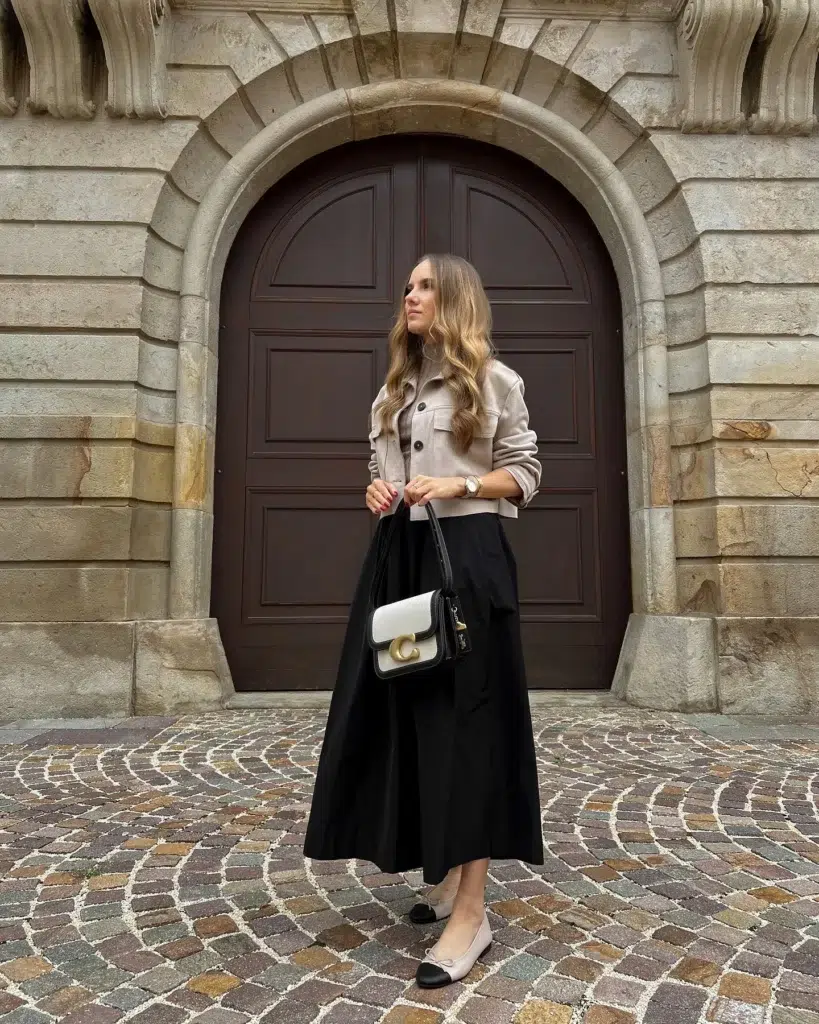 Petite woman in a cropped sand beige utility-style jacket with patch chest pockets and a button front, worn over a high-waisted full flared black midi skirt reaching mid-calf, paired with two-tone cap-toe ballet flats in nude with a black toe cap, carrying a structured two-tone black and cream crossbody bag with a large gold hardware clasp, wearing a gold watch, standing on a cobblestone courtyard in front of a grand dark arched wooden doorway set in ornate stone architecture.