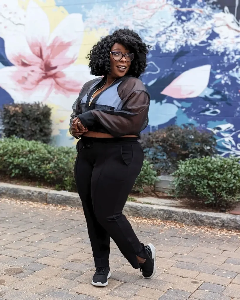 Curvy petite woman standing on a cobblestone street in front of a large painted floral mural, wearing a sheer dark cropped bomber jacket with a gold zipper over a periwinkle blue crop top, paired with black high-waist tapered pants and black athletic sneakers with white soles, wearing large cat-eye glasses and natural voluminous curly hair worn loose.
