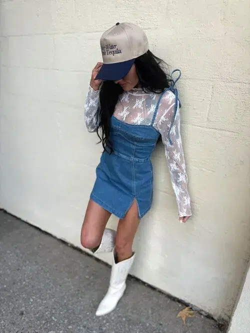 Woman in a medium blue wash fitted denim mini pinafore dress with thin adjustable bow-tie shoulder straps, a structured corset-style bodice, and a small front hem slit, layered over a white sheer floral lace long-sleeve top with sleeves extending past the denim, styled with a tan beige trucker-style baseball cap with embroidered text, and white pointed-toe cowboy boots with a low stacked heel, leaning against a white painted exterior wall in an outdoor street setting.