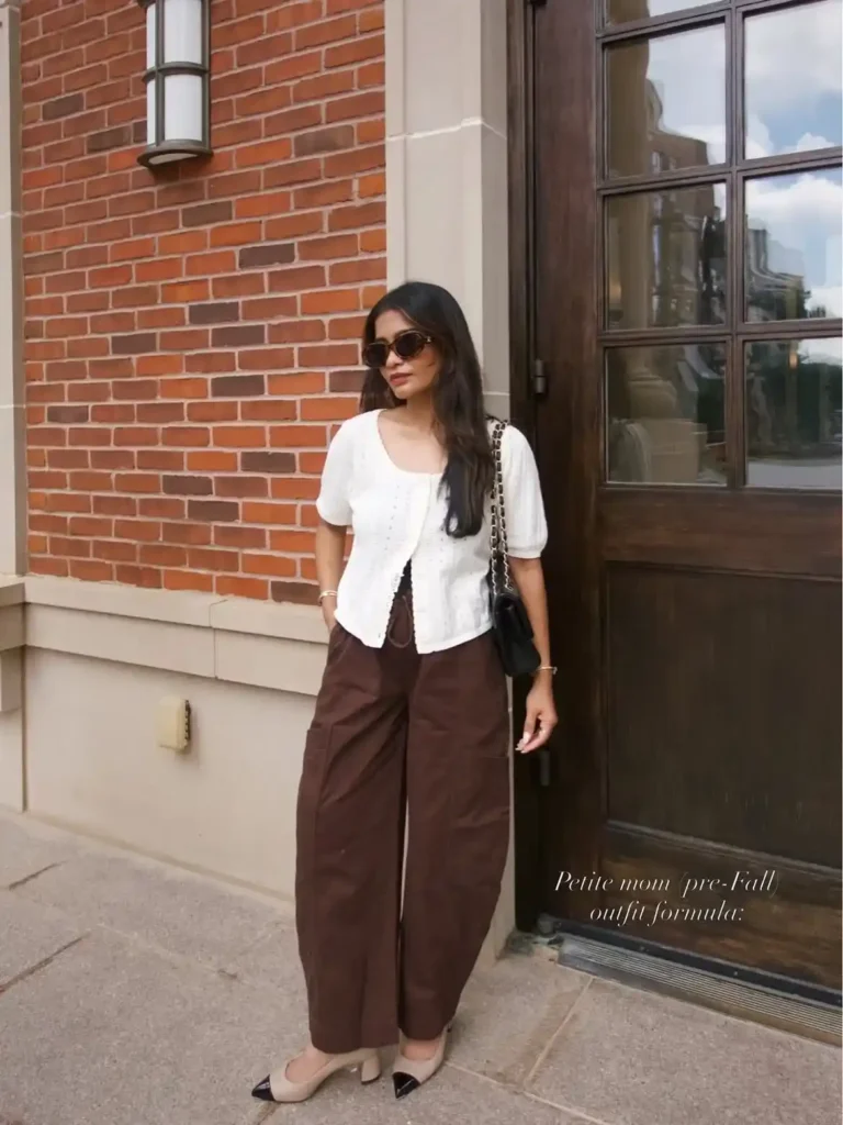 A petite woman standing outside a red brick building beside a large dark wooden door, wearing a white short-sleeve textured eyelet open-front cardigan over a white camisole, paired with dark chocolate brown relaxed wide-leg trousers with side patch pockets, nude cap-toe block-heel pumps, and a small black quilted chain-strap shoulder bag, with tortoiseshell sunglasses.