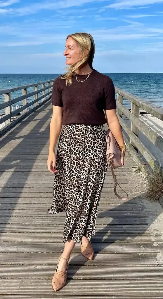 A woman walking along a wooden coastal boardwalk on a sunny day with blue sea and sky behind her, wearing a dark oxblood brown short-sleeve fine knit sweater tucked softly over the waistband of a fluid leopard print satin midi skirt in cream, black, and brown tones falling to mid-calf, paired with nude pointed-toe heeled mules and a blush pink structured handbag with layered gold necklaces.