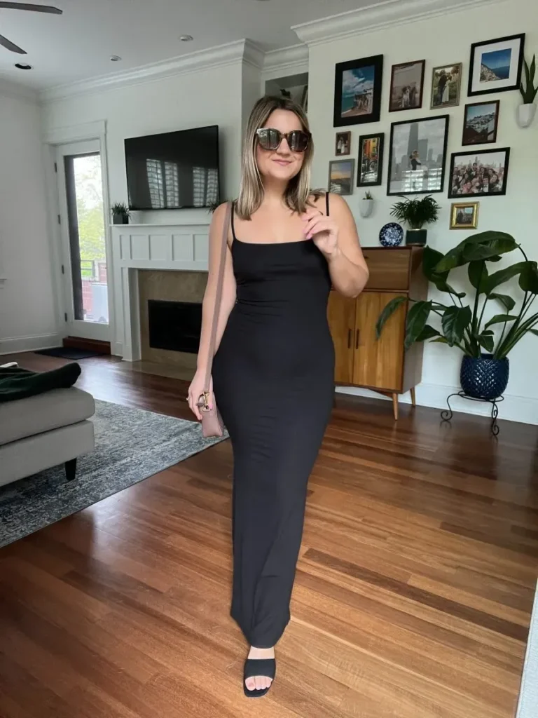 Woman with shoulder-length blonde hair wearing a fitted black maxi slip dress with wide square straps and a scoop neckline in matte fabric, paired with black flat slide sandals, a small dusty blush pink clutch with gold hardware, and large tortoiseshell cat-eye sunglasses, standing in a bright modern living room with hardwood floors, a gallery wall, and a mid-century sideboard.