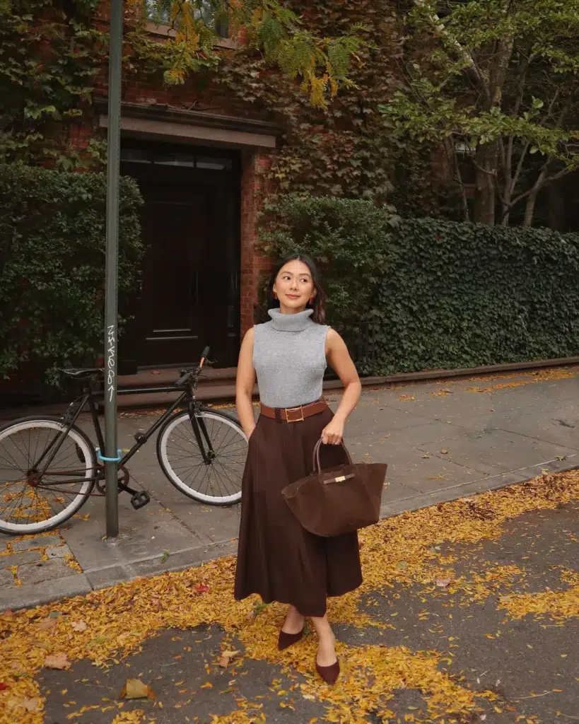 Petite woman in a sleeveless gray ribbed turtleneck tucked into a deep chocolate brown full midi skirt, cinched with a tan leather belt and gold buckle, carrying a deep brown suede structured tote bag with gold hardware, wearing dark brown pointed-toe mules, standing on a city sidewalk blanketed in fallen yellow autumn leaves with an ivy-covered brick building behind her.