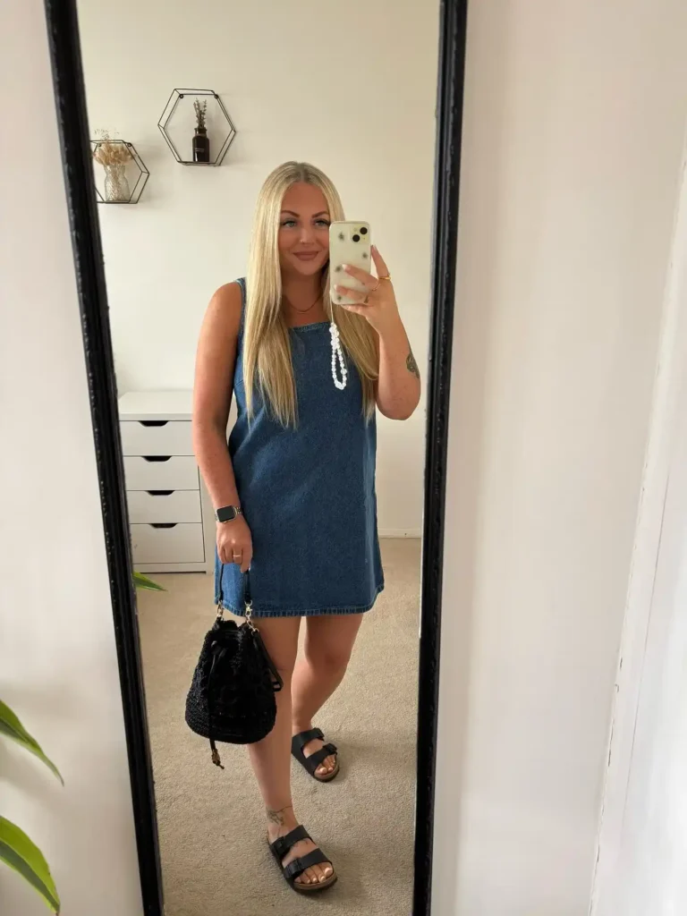 Woman taking a mirror selfie in a medium blue wash sleeveless square-neck denim shift mini dress with a straight clean hem and visible topstitching, styled with a chunky white beaded necklace, a black woven crochet drawstring bucket bag with a leather and chain handle, black double-buckle footbed sandals, a black smartwatch, and a simple gold ring, photographed in a minimal white bedroom with a black-framed full-length mirror, white drawer unit, and small geometric hexagonal wall shelves.