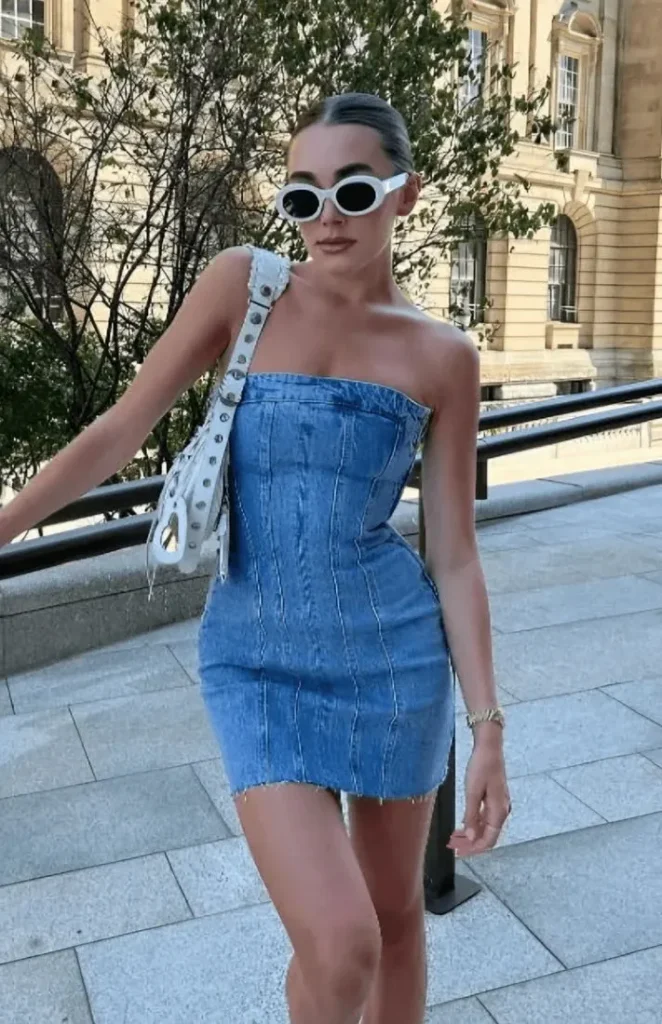 Woman in a medium blue wash strapless fitted denim mini dress with visible vertical corset-style seam lines throughout the bodice and a raw-cut hem, styled with white oval retro sunglasses, a white structured shoulder bag with a grommet-detail strap, a gold bracelet, and a slicked-back bun hairstyle, standing on stone pavement steps near a metal railing with a classical stone building in the background.